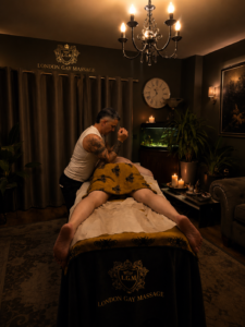 Massage therapist working on a client lying face down on a padded table in a candlelit, upscale room labeled London Gay Massage (LGM).